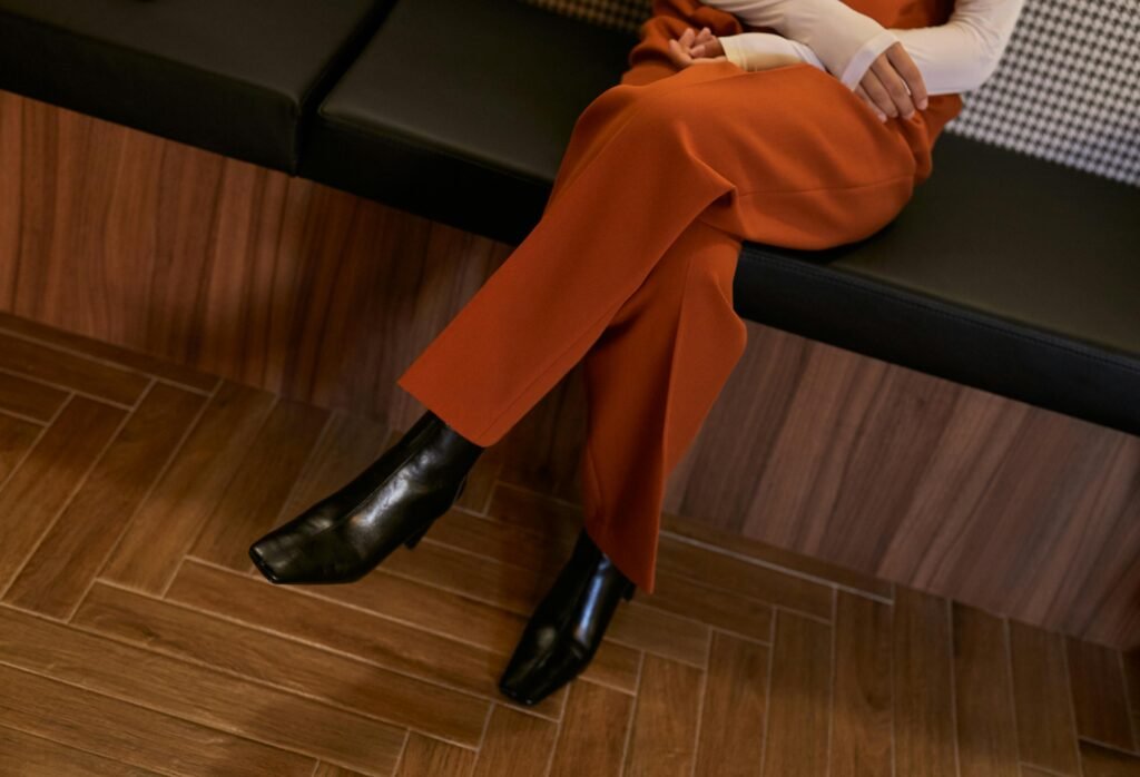 pexels-photo-10339245-10339245 Low-angle view of an unrecognizable woman in orange pants and elegant black boots sitting with legs crossed indoors.